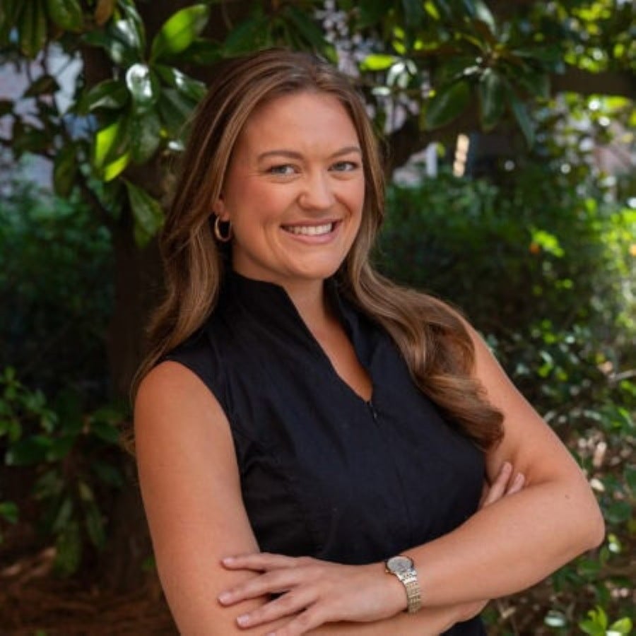 Professional outdoor headshot of Dr. Braidyn Lazenby, a woman with long, wavy brown hair, wearing a sleeveless black high-neck top, gold hoop earrings, and a watch. She is smiling cheerfully at the camera with her arms crossed, posed in front of lush green foliage and trees.