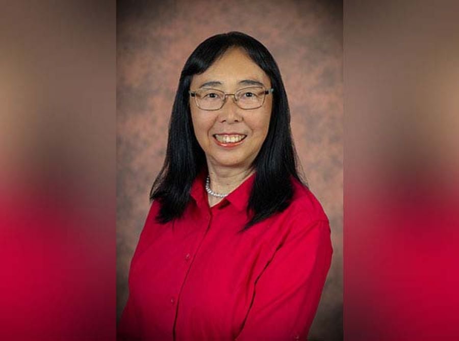 Professional headshot of Dr. Qing-Xiang “Amy” Sang, a woman with dark, shoulder-length hair and straight bangs, wearing wire-rimmed glasses and smiling warmly at the camera. She is wearing a vibrant red button-up shirt and a white pearl necklace, posed against a textured, neutral studio backdrop.