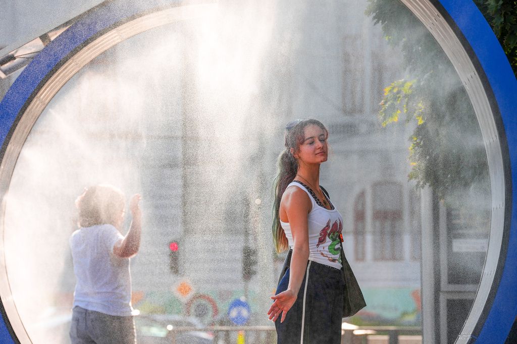 A young woman in a white tank top closes her eyes to cool off inside a large circular misting station on a sunny city street. Fine water mist glows in the sunlight around her as another pedestrian walks through the misting arch, with blurred historic buildings in the background.