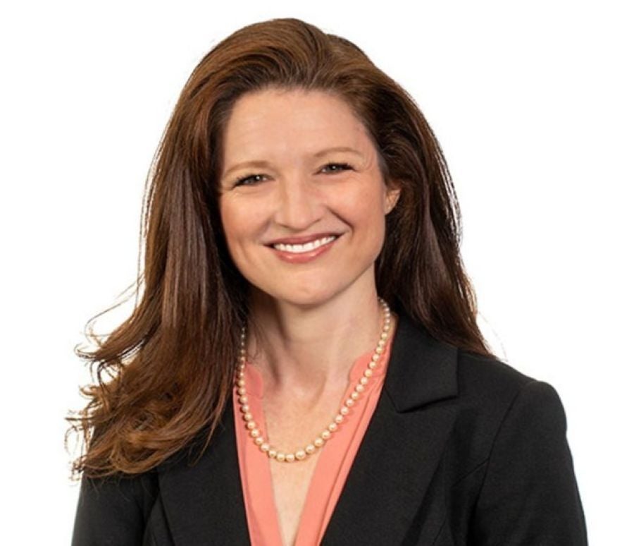 Professional headshot of Dr. Dawn Carr smiling against a plain white background. She has long, wavy reddish-brown hair draped over her right shoulder and is wearing a black blazer over a peach-pink V-neck top, styled with a single-strand cream pearl necklace.
