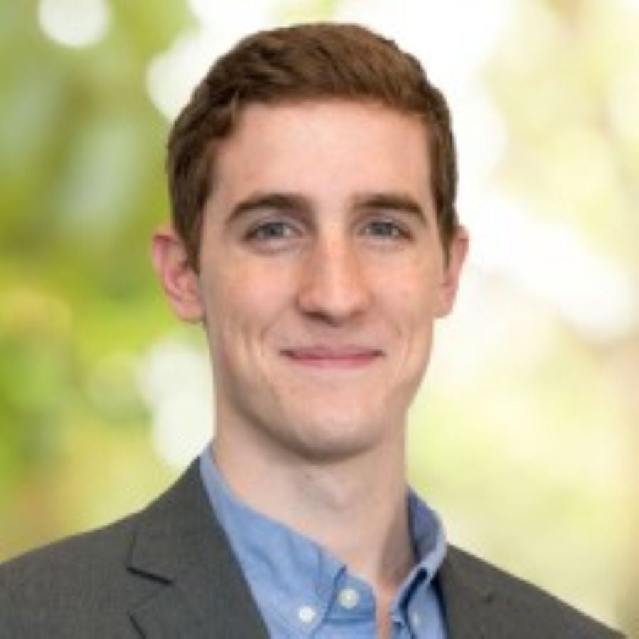 Headshot of Joseph Watso, a man with short brown hair and light eyes, smiling gently at the camera against a softly blurred green and cream background. He is wearing a dark grey suit jacket over a light blue button-up shirt with an open collar.