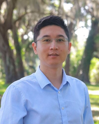 Headshot of Guang Wang wearing glasses and a light blue shirt outdoors.