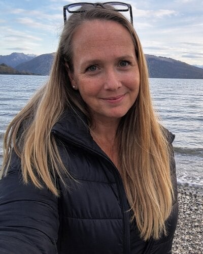 a woman with long, straight blonde hair smiling warmly at the camera. She wears a black puffer jacket with glasses resting on her head, standing on a rocky beach next to a wide lake. The background features a scenic view of distant snow-covered mountains under a soft, overcast sky.