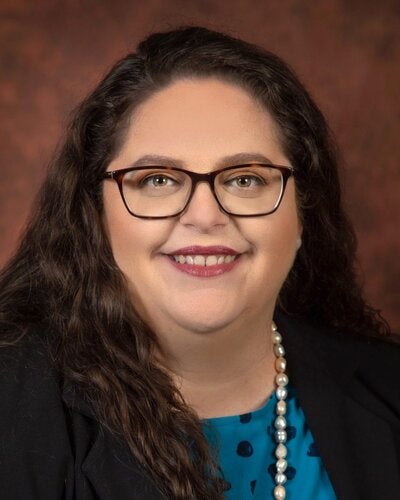 A professional studio headshot of a woman with long, dark wavy hair framing her face. She is smiling warmly at the camera, wearing rectangular tortoiseshell glasses and lipstick. She is dressed in a black blazer layered over a bright teal and blue polka-dot top, accented by a single strand pearl necklace. The background is a softly blurred, textured reddish-brown studio backdrop.