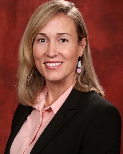 Professional headshot of Ursula Weiss, smiling and wearing a black blazer over a light pink collared shirt, set against a warm reddish-brown background.