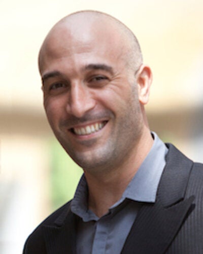 A close-up headshot of a smiling bald man with brown eyes, dark eyebrows, and light stubble along his jawline. He is wearing a dark greyish-black suit jacket over a light grey collared button-up shirt, posed against a soft, warm outdoor background.