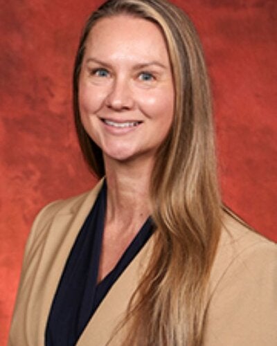 Headshot of Heather Robinson, a woman with long, straight light-brown hair styled over her left shoulder. She is smiling at the camera, wearing a dark navy top and a light tan or beige blazer, set against a textured dark red background.   