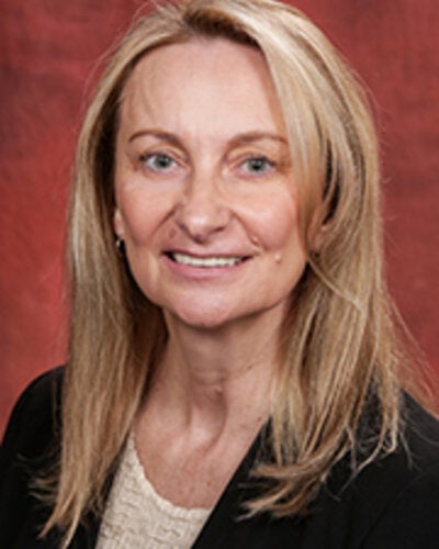 Headshot of Ewa Bienkiewicz, a woman with blonde, straight, shoulder-length hair parted down the middle. She is smiling gently at the camera, wearing a dark jacket or blazer over a light-colored textured top, set against a dark red backdrop.