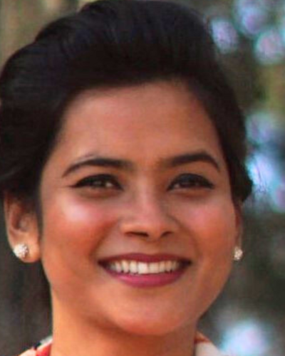 Close-up portrait of Tania Sultana smiling brightly. She has dark hair styled in an updo, wears stud earrings, and is set against a softly blurred background with natural lighting.   