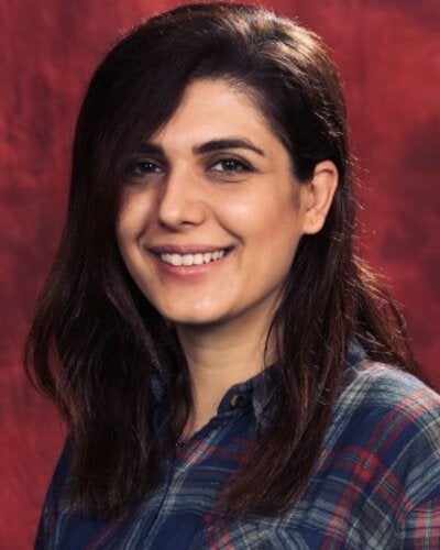 A headshot portrait of nina zamani with dark shoulder-length hair and a side part. She is smiling warmly at the camera and wearing a blue, red, and grey plaid button-down shirt against a mottled dark red backdrop.