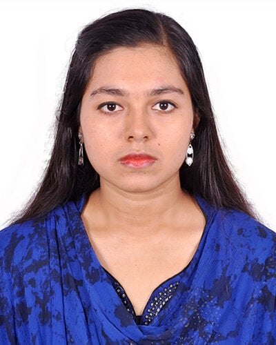 A studio headshot portrait of Farzana Mim against a plain, off-white background. She has long, straight dark hair worn loose with a center part, tucked behind her ears to show small white hoop or bead earrings. She faces directly forward with a calm, neutral facial expression. She is wearing a vibrant royal blue top with a scoop neckline and subtle dark pattern detailing.   